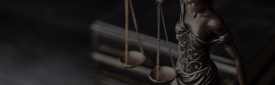 A dark, professional image featuring a bronze Lady Justice statue holding scales. The background is blurred.